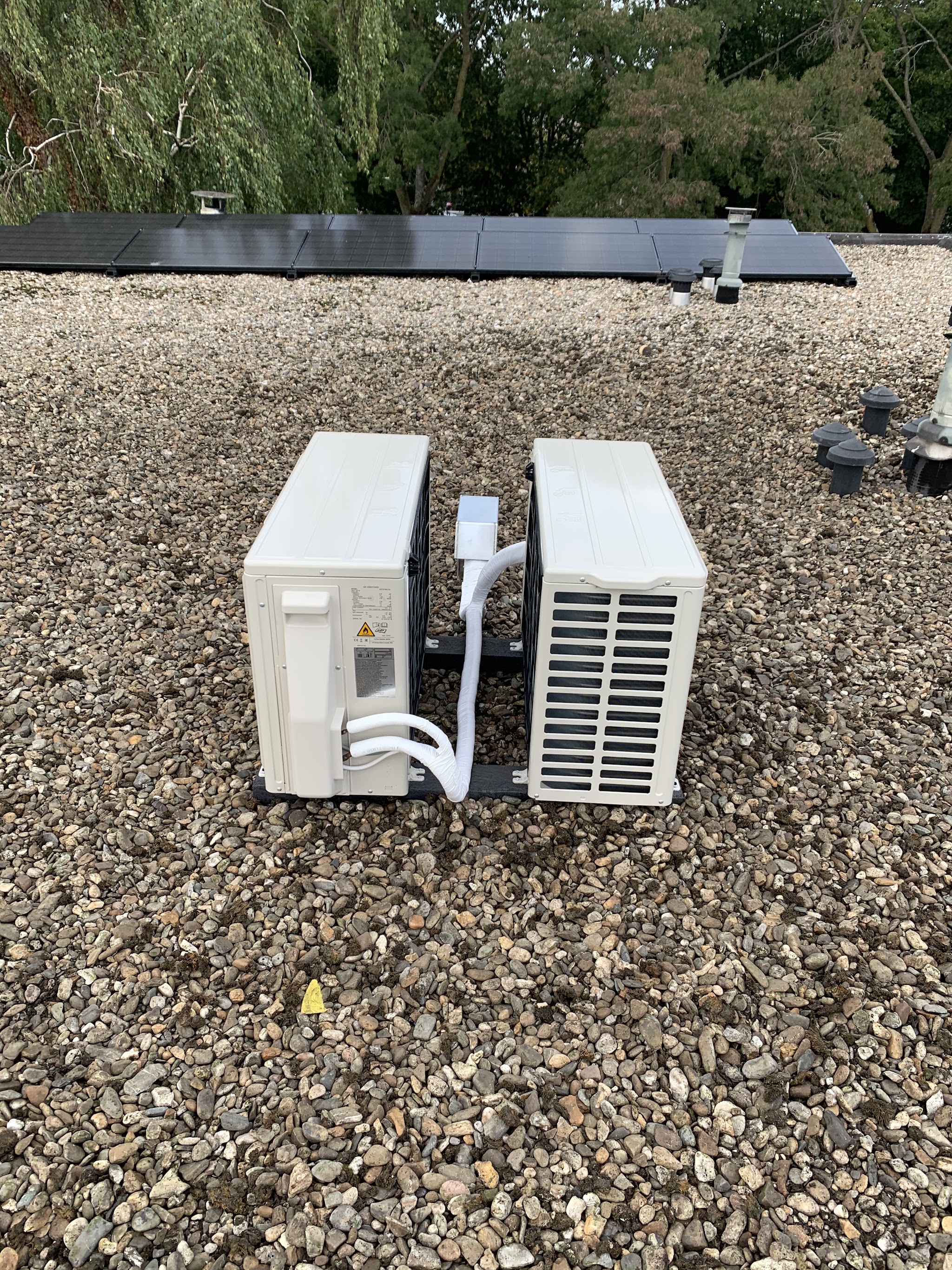 Twee buitenunits naast elkaar op een grinddak, zonnepanelen op de achtergrond
