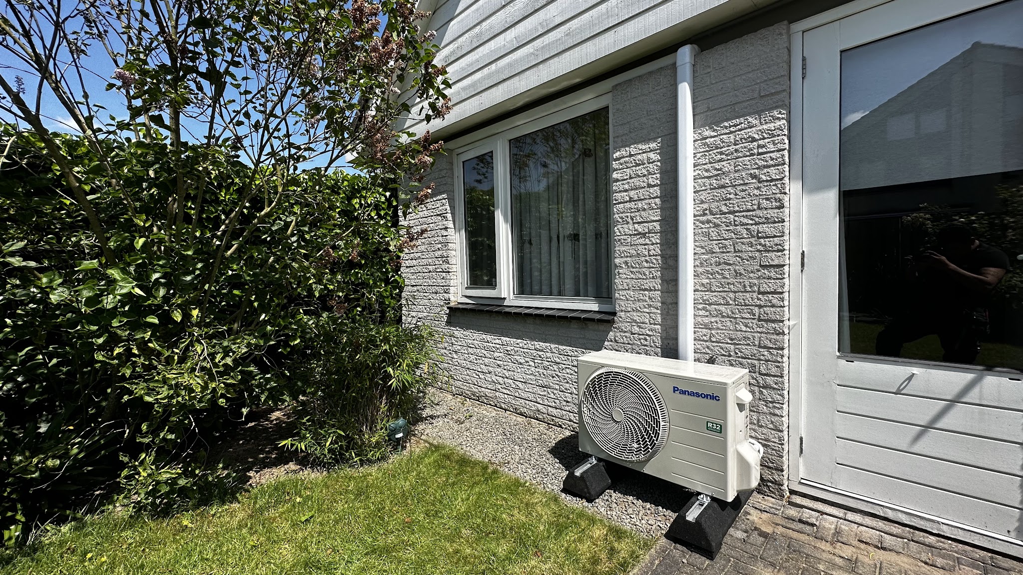 Panasonic buitenunit op pootjes in een achtertuin, leidingen netjes in een witte goot