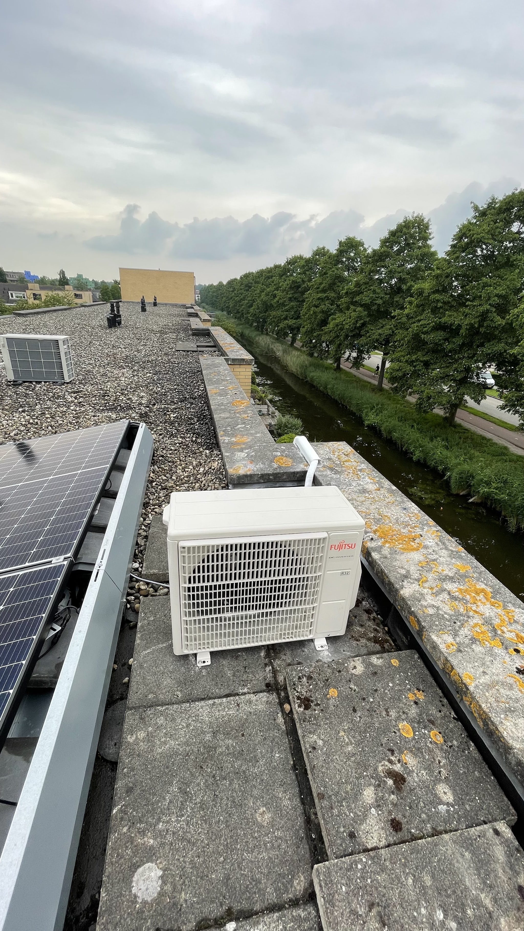 Buitenunit op de rand van een plat dak naast zonnepanelen, met uitzicht over een vaart
