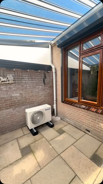 Panasonic buitenunit onder een glazen veranda naast een bakstenen muur